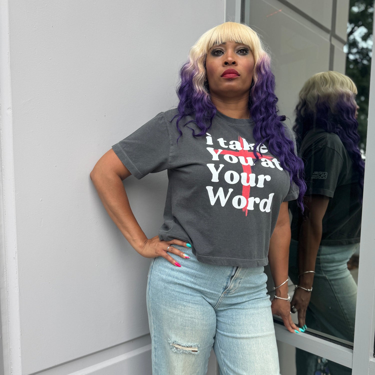Person wearing a gray t-shirt with text and light blue jeans standing against a light-colored wall.